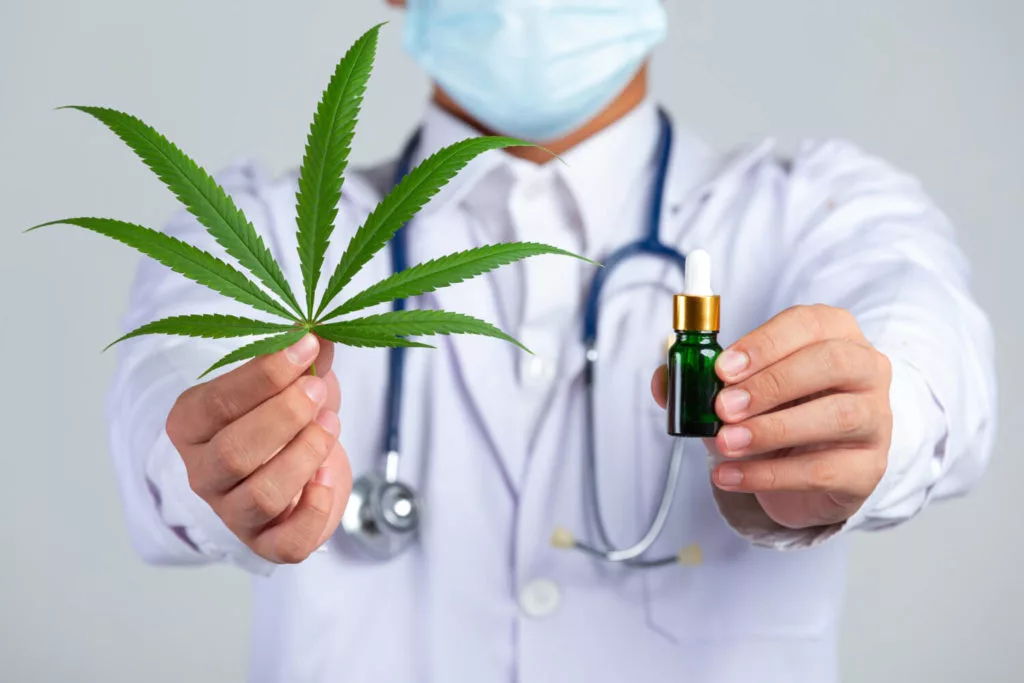 medical doctor holding cannabis leaf and bottle of cannabis oil on white wall 1 1024x683 1 jpg